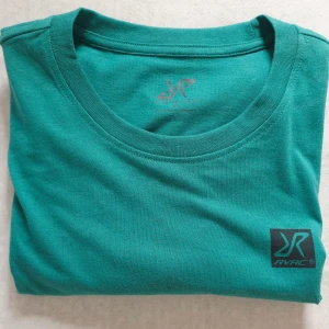 Turkos t-shirt från RVRC i bomull - Snygg turkos t-shirt från RVRC med rund hals och korta ärmar. T-shirten har en diskret svart RVRC-logga på bröstet och en liten etikett i sidan. Tillverkad i mjuk bomull som känns skön mot huden. Perfekt för dig som gillar stilrena plagg med enkel design.