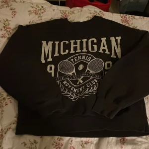 Svart Michigan sweatshirt Gina Tricot XS - Svart sweatshirt från Gina Tricot i storlek XS med stort Michigan-tryck och tennisracketar framtill. Tröjan har ribbade muddar vid hals, ärmslut och nederkant. Mjuk fleece på insidan för extra komfort. Perfekt för dig som gillar sportig och college-inspirerad stil.