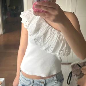 Vit oneshoulder topp med spetsvolang - Säljer en vit oneshoulder topp med bred spetsvolang runt halsen. Volangen har broderade blommönster och ger en romantisk vibe. Toppen är croppad och ärmlös, perfekt till ljusa jeans eller kjol. Materialet känns som mjuk bomull.