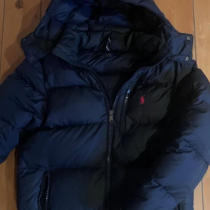 Mörkblå dunjacka Polo Ralph Lauren - Snygg mörkblå dunjacka från Polo Ralph Lauren med huva och dragkedja. Jackan har klassisk pufferstil, lång ärm och ikonisk röd logga på bröstet. Perfekt för kalla dagar och riktigt skön att bära. Fodrad och med praktiska fickor. (VIKTIGT —>) båda fickorna ska egentligen ha dragkedjor detaljen men båda har gått sönder som man ser på bilden, du kan fortfarande stänga och öppna fickorna men det blir svårare pga detta. Det finns ett litet hål i luvan som inte syns när man använder jackan och kan sys