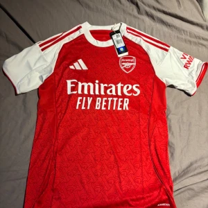 Arsenal hemmatröja Adidas röd/vit S - Arsenal hemmatröja från Adidas i rött med vita ärmar och detaljer. Tröjan har korta ärmar, rund hals och ett subtilt mönster över hela framsidan. Materialet är lätt och andas, perfekt för fotboll eller att bära till vardags. Klassisk klubbsköld och sponsortryck på bröstet.