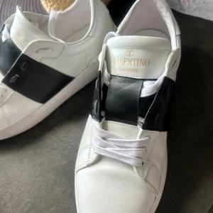 Valentino Garavani vita sneakers - Snygga vita sneakers från Valentino Garavani med bred svart läderdetalj över snörningen och ikoniska nitar på hälen. Skorna har rund tå, platt sula och är tillverkade i mjukt skinn. Perfekt för dig som gillar exklusiv och stilren design.