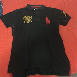 Svart piké från Polo Ralph Lauren - Svart pikétröja från Polo Ralph Lauren i custom fit med klassisk krage och korta ärmar. Stora broderade detaljer i rött och gult framtill samt broderad siffra på ärmen. Tillverkad i mjuk bomull, perfekt för en clean och sportig look. Jag säljer den på grund av att den är för liten och fick den i present och it går att skicka tillbaks 