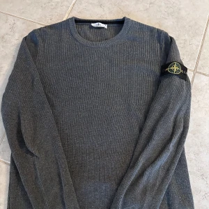 Grå stickad tröja Stone Island - Tröjan är bara använd fåtal gånger. Snygg grå stickad tröja från Stone Island med klassisk rund hals och ribbad struktur. Tröjan har lång ärm och den ikoniska Stone Island-patchen på vänster arm. Perfekt för dig som gillar stilrena och tidlösa plagg med street-vibe.