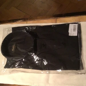 Svart skjorta från Casino Cosmopol - Stilren svart skjorta från Casino Cosmopol i klassisk modell med knappar framtill och snygg krage. Perfekt för dig som gillar en clean och tidlös look. Skjortan är i storlek 41/42 och har en enkel, enfärgad design.