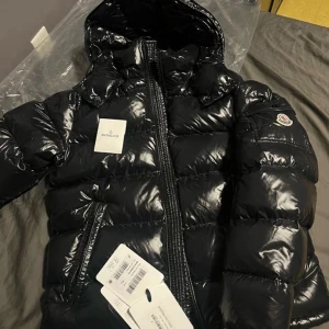 Svart pufferjacka från Moncler - Säljer en svart pufferjacka från Moncler med glansig finish och huva. Jackan har lång ärm, dragkedja framtill och klassisk Moncler-logga på ärmen. Perfekt för kalla dagar och riktigt snygg streetstil.