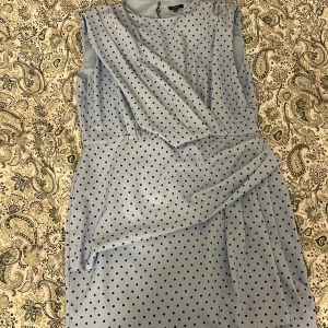 Ljusblå prickig klänning Floryday - Supersöt ljusblå klänning från Floryday med svarta prickar och omlottdetalj framtill. Klänningen är ärmlös och har en elegant, mjuk form som ger en snygg siluett. Perfekt för dig som gillar stilrena mönster och en feminin look.