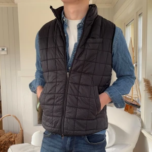 Svart quiltad dunväst John Henric M - Svart dunväst från John Henric i storlek M. Västen har ett quiltat rutmönster, hög krage och dragkedja framtill. Perfekt att slänga över en hoodie eller skjorta för extra värme. Snygg och enkel design som funkar till jeans eller chinos.