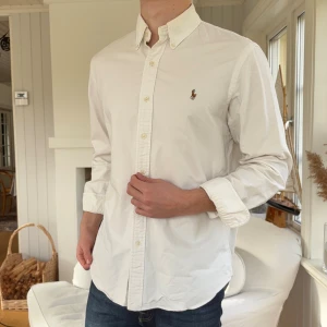 Vit skjorta från Polo Ralph Lauren - Vit skjorta från Polo Ralph Lauren med klassisk button-down krage och broderad logga på bröstet. Skjortan har lång ärm, knappar framtill och är tillverkad i mjuk bomull. Perfekt för dig som gillar stilrena och tidlösa plagg.