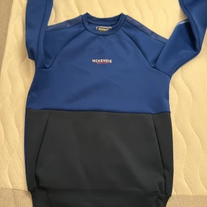 Blå och mörkblå sweatshirt från McKenzie - Säljer en snygg tvåfärgad sweatshirt från McKenzie i blått och mörkblått. Tröjan har lång ärm, rund hals och logga på bröstet samt tryckt McKenzie-text längs ärmarna. Tillverkad i mjukt syntetmaterial, perfekt för en sportig och avslappnad stil.
