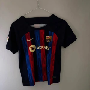 FC Barcelona matchtröja Nike blå/röd - Säljer en FC Barcelona matchtröja från Nike med klassiska blå och röda ränder, guldiga detaljer och klubbmärke. Tröjan har korta ärmar, La Liga-märke på ärmen och tryck med nummer 10 på ryggen. Materialet är lätt och andas, perfekt för fotboll eller häng.