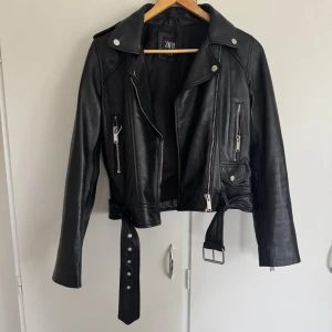 Svart skinnjacka från Zara - Snygg svart skinnjacka från Zara med klassisk bikerstil. Jackan har flera silvriga dragkedjor, knappar och ett bälte i midjan. Croppad passform och långa ärmar. Perfekt för dig som vill ha en edgy look med coola detaljer. ÄKTA SKINN!! Lite liten i storlek, sitter mer som en S