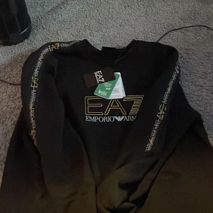 Svart EA7 Emporio Armani sweatshirt - Svart sweatshirt från EA7 Emporio Armani med guld- och vitfärgad logga på bröstet och tryck längs ärmarna. Klassisk rund hals och långärmad modell. Perfekt för dig som gillar stilrena och exklusiva plagg med streetkänsla.