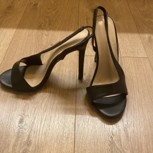 Svarta pumps med öppen tå och klack - Snygga svarta pumps med öppen tå och hög klack. Modellen har en stilren design med smal rem runt hälen och bred rem över foten. Skorna har en rundad framdel och är tillverkade i ett slätt material som ger en elegant look.