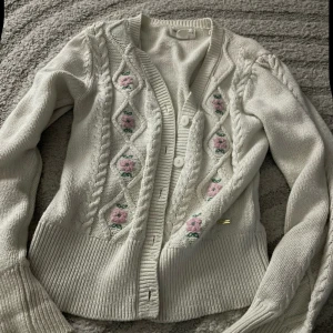 Vit kofta med broderade blommor Holly & Whyte - Superfin vit kofta från Holly & Whyte med broderade rosa blommor och gröna blad framtill. Tröjan har kabelstickade detaljer, ribbad nederkant och muddar, samt knappar framtill.