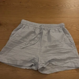 Randiga ljusblå shorts från Pigalle - Säljer ett par fräscha ljusblå och vita randiga shorts från Pigalle i storlek S. De har elastisk midja med snörning och är gjorda i mjuk bomull. Perfekta för chill dagar och har en loose passform som ger skön vibe.