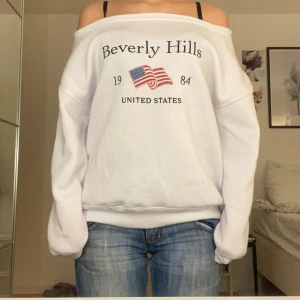 Vit offshoulder Beverly Hills sweatshirt - Vit sweatshirt med offshoulder-design och tryck framtill med texten 'Beverly Hills', amerikansk flagga och årtal 1984. Tröjan har lång ärm och bred halsringning som ger en chill vibe. Mjuk och skön, perfekt till jeans eller shorts.