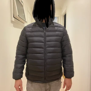 Svart pufferjacka med huva - Säljer en svart pufferjacka med huva och dragkedja framtill. Jackan har horisontella sömmar, lång ärm och en clean, enkel look. Perfekt för kalla dagar och riktigt skön att bära. Passformen är normal och jackan är vadderad för extra värme. Helt ny 