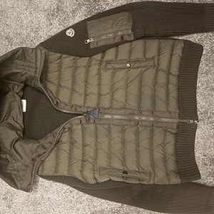 Moncler mörkgrön dunjacka med huva - Snygg mörkgrön dunjacka från Moncler med stickade partier och quiltad framsida. Jackan har huva, dragkedja, fickor med knapp och en extra ficka med dragkedja på ärmen. Perfekt för dig som vill ha en cool och varm jacka med streetkänsla. Nackdel med jackan är att det lite smuts på moncler märket annars bra 