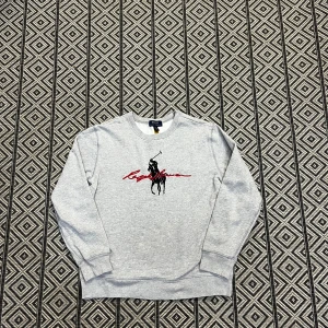 Grå sweatshirt från Polo Ralph Lauren - Snygg grå sweatshirt från Polo Ralph Lauren med klassiskt svart polospelartryck och röd signatur på bröstet. Tröjan har rund hals, ribbade muddar och är tillverkad i mjuk bomullsmix. Perfekt för en chill och stilren look. Om ni har frågor är det vara stt höra av er. Skickas samma dag!