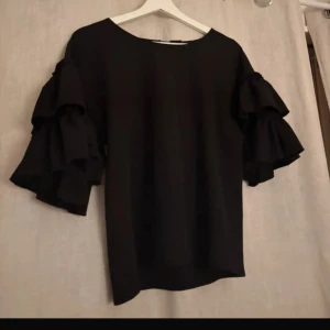 Svart blus med volangärmar från Gina Tricot - Säljer en svart blus från Gina Tricot med bred rund halsringning och korta ärmar med flera lager volanger. Toppens material känns mjukt och lätt, perfekt för dig som vill ha en stilren men ändå extra look. Passar till många olika outfits!