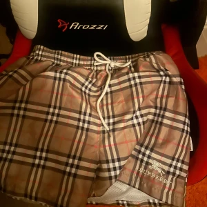 Rutig Burberry shorts XL med snörning - Säljer ett par klassiska Burberry badshorts/shorts i beige med rutigt mönster och vita, svarta och röda detaljer. Shortsen har snörning i midjan och innerbyxa i mesh. Perfekta för sommaren och riktigt snygga till streetwear-stilen. Skulle säga att de är ungefär 170-180. 