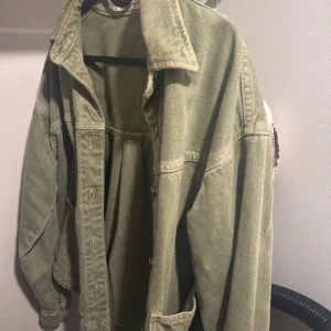 Olivgrön jeansjacka med knappar - Snygg olivgrön jeansjacka med klassisk krage och stora fickor framtill. Jackan har knappar längs hela framsidan och är i ett lite loose fit. Perfekt att slänga över en hoodie eller t-shirt för en chill look.