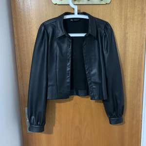 Svart skinnjacka från Zara, S - Snygg svart skinnjacka från Zara i storlek S. Jackan är croppad med klassisk krage, knappar framtill och långa ärmar med lätt puff vid axlarna. Perfekt för dig som vill ha en stilren och trendig look.