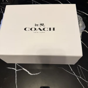 Svart axelväska från Coach i skinn - Snygg svart axelväska från Coach i äkta skinn med guldig C-detalj framtill. Stilren och klassisk design med lock och medföljande axelremmar. Perfekt för dig som vill ha en tidlös och trendig väska med exklusiv känsla.