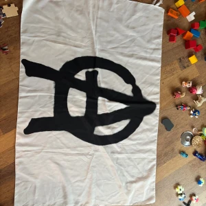 Vit anarki flagga  med svart tryck - Flagga Anarki i vitt tyg med svart handmålat tryck som syns på både fram- och baksidan. Flaggan har knytband i sidorna för att fästas 