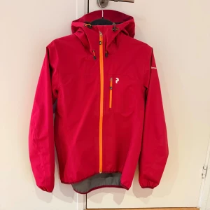 Röd rosa vindjacka Peak Performance S - Snygg röd rosa vindjacka från Peak Performance med huva och orange dragkedja. Jackan har en bröstficka med dragkedja, elastiska muddar och diskreta loggor på bröstet och huvan. Perfekt för blåsiga dagar och outdoor-aktiviteter. Skicket på jackan är mycket bra men använd och storleken är S.