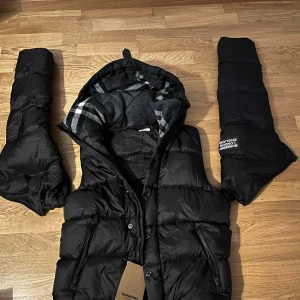 Burberry Jacka Puffer - Säljer en svart pufferjacka från Burberry som har avtagbara ärmar med klassiskt rutigt foder. Jackan har dragkedja, tryckknappar och flera fickor med dragkedja. Stort Burberry-märke på ena ärmen. Perfekt för kalla dagar och riktigt snygg streetstil.