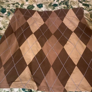 Brun rutig kortkjol i bomull - Snygg kortkjol med rutigt argylemönster i olika bruna nyanser. Kjolen är gjord i mjuk bomull och har en klassisk, rak passform. Perfekt för dig som gillar preppy stil och vill sticka ut lite extra.