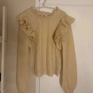 Beige stickad tröja med volang JDY - Superfin beige stickad tröja från JDY i storlek S. Tröjan har snygga volangdetaljer på axlarna och ett klassiskt kabelstickat mönster framtill. Mjuk och skön med rund hals och långa ärmar, perfekt för att hålla sig varm och ändå se stylish ut.
