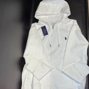 Vit hoodie med en klassisk svart logga - Snygg vit zip-hoodie med dragkedja och huva. Klassisk svart logga broderad på bröstet och praktiska fickor framtill. Tillverkad i mjuk bomull för en skön känsla. Perfekt för en avslappnad och stilren look.