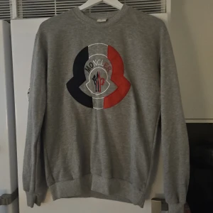 Grå sweatshirt - Säljer en grå sweatshirt i storlek XL. Tröjan har ett stort broderat logo på bröstet i svart, vitt och rött. Klassisk rund halsringning och ribbade muddar vid ärmslut och nederkant. Perfekt för dig som gillar stilrena märkesplagg.
