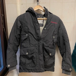 Svart parkajacka från Didriksons 150 - Svart parkajacka från Didriksons med huva och fuskpäls, flera stora fickor med knappar och dragkedja. Jackan är fodrad med mjuk teddy på insidan och har vattentätt Stormsystem-tyg. Perfekt för kalla och blöta dagar.