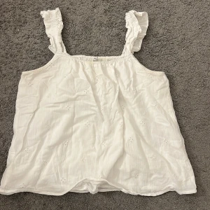 Vit blommig linnetopp från ONLY - Supersöt vit linnetopp från ONLY med broderade små blommor och volangprydda axelband. Toppen är ärmlös och har en luftig passform, perfekt för varma dagar. Materialet känns mjukt och lätt mot huden. Säljes i storlek M!