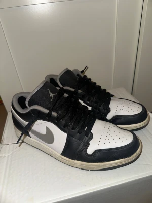 Nike Air Jordan 1 Low svart/vit - Nike Air Jordan 1 Low sneakers i svart och vitt med grå detaljer. Klassisk siluett med snörning, rund tå och platt sula. Skorna har Air Jordan-logga broderad på hälen och Swoosh på sidan. Tillverkade i skinn och syntetmaterial för en clean look. Det sitter laglappar i båda hälarna, ingenting som känns. 