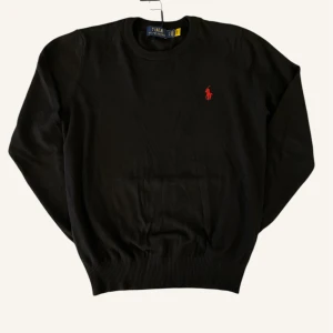 Svart tröja från Polo Ralph Lauren - Svart tröja från Polo Ralph Lauren med klassisk rund hals och rött broderat logomärke på bröstet. Tröjan har ribbade muddar vid ärmslut och nederkant, och är tillverkad i mjuk bomull som känns skön mot huden. Perfekt för dig som gillar stilrena och tidlösa plagg.