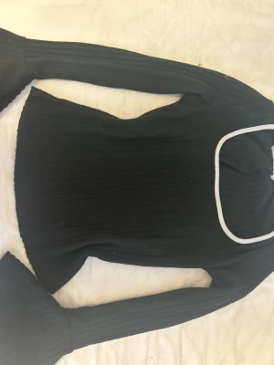 Svart ribbad långärmad topp med vit kant - Svart ribbad topp med lång ärm och vit kant runt den fyrkantiga halsringningen. Modellen är figurnära med utsvängda ärmslut och har en cool, modern vibe. Perfekt för dig som gillar stilrena detaljer och vill sticka ut med en enkel men snygg look.