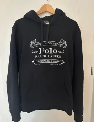 Svart hoodie Polo Ralph Lauren  - Svart hoodie från Polo Ralph Lauren i storlek XL med vit tryckt logga och text på bröstet. Tröjan har huva med snörning, stor magficka och långa ärmar. Mjuk och skön bomullsmix som passar perfekt för chill dagar. Snygg streetstil och klassisk design.