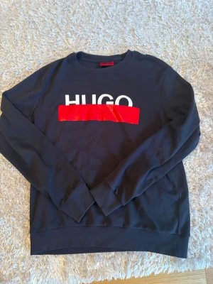 Svart sweatshirt från Hugo Boss - Snygg svart sweatshirt från Hugo Boss med stort HUGO-tryck i vitt och rött framtill.  Använt ett par gånger, skick 8/10. Tröjan är äkta  såklart  