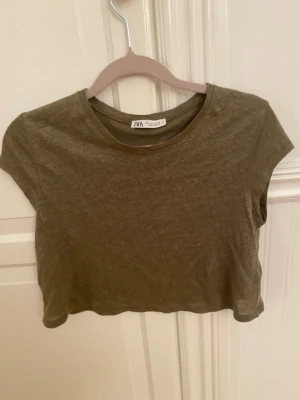 Olivgrön croppad topp från Zara - Säljer en olivgrön croppad topp från Zara i storlek S. Toppen har rund halsringning och korta ärmar, tillverkad i mjuk bomull. Perfekt att styla med höga jeans eller shorts för en chill look. Enkel och stilren design utan mönster eller detaljer.
