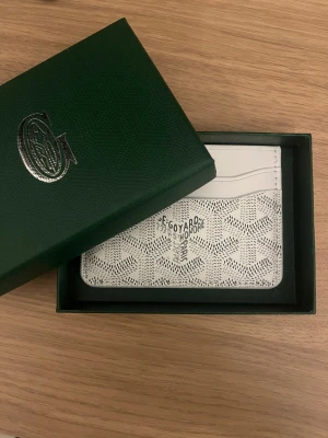 Vit Goyaard plånbok med mönster - Snygg vit plånbok från Goyaard med klassiskt grafiskt mönster och flera kortfack. Plånboken är tillverkad i skinn och har Goyaard Paris-logga präglad på framsidan. Perfekt för dig som vill ha en stilren och exklusiv accessoar.