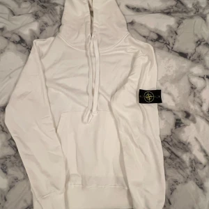 Vit hoodie från Stone Island - Vit hoodie från Stone Island med klassisk patch på vänster ärm. Tröjan har huva med snörning, stor magficka och långa ärmar. Mjuk bomullskvalitet och stilren design som ger en clean look. Perfekt för dig som gillar streetwear och vill ha något snyggt och bekvämt.