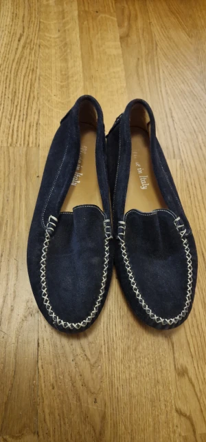 Mörkblå mocka loafers Made in Italy - Snygga mörkblå loafers i mjuk mocka med kontrasterande vit söm runt tån. Klassisk rund tå och platt sula med gummidetaljer bak för extra grepp. Perfekt för dig som gillar stilrena och bekväma skor med italiensk touch.