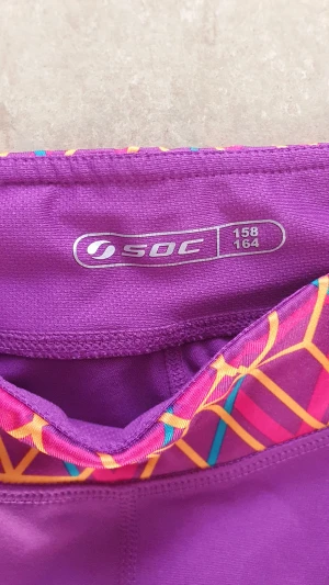 Ankellånga träningsleggings - Snygga lila ankellånga träningsleggings från SOC i storlek 158/164. De har ett färgglatt geometriskt mönster i midjan med inslag av orange, rosa och blått. Modellen är trekvarts och materialet är syntet, perfekt för aktiva dagar.