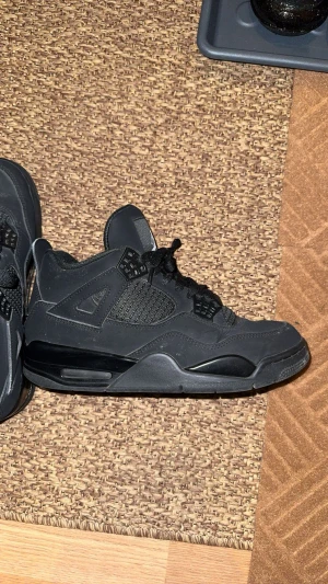 Svarta Nike Jordan 4 sneakers - Säljer ett par svarta Nike Jordan 4 sneakers med klassisk meshpanel, snörning och Jumpman-logga på plösen och yttersulan. Skorna har en chunky siluett, platt sula och detaljer i syntet och mocka för en cool streetstil. Perfekt för dig som gillar retro och sportig vibe.