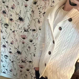 Vit kabelstickad kofta med knappar - Säljer en vit kabelstickad kofta med grov krage och stora bruna knappar. Koftan har ett klassiskt flätmönster och är långärmad, perfekt för att hålla sig varm och snygg under kyliga dagar. Passar dig som gillar mysiga och stilrena plagg.